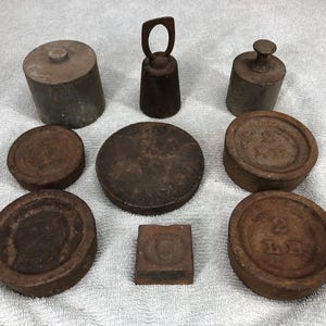 May include: An assortment of antique metal weights in various shapes and sizes. The collection includes cylindrical, round, and square weights, with a bell-shaped weight with a handle. The weights are a rusty brown color.