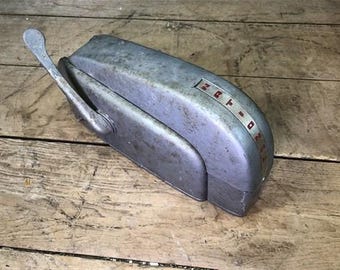 Puede incluir: Un objeto metálico gris desgastado con una palanca y la palabra "Zeta" en letras rojas. El objeto de diseño curvo parece estar hecho de metal resistente. El artículo está sobre un fondo de madera.