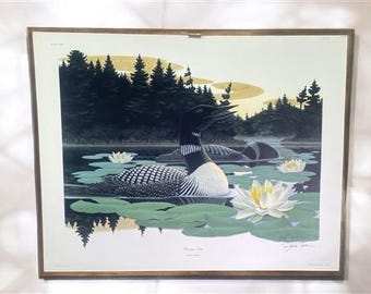 May include: A framed print of a black and white loon swimming in a lake with lily pads and yellow flowers. The print is signed by the artist, "Sylvia  Slater".