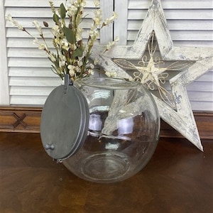 Pode incluir: Um frasco de vidro transparente com uma tampa de metal. O frasco é redondo e tem uma boca larga. A tampa é prateada e tem uma pega no topo. O frasco está sentado numa mesa de madeira.