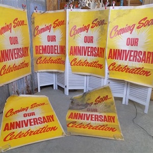 May include: Five yellow and red paper signs with the text "Coming Soon... Our Anniversary Celebration" and "Coming Soon... Our Remodeling Celebration". The signs are propped up on a white wooden screen.