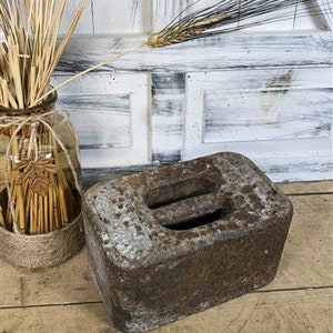 May include: A weathered, rectangular metal weight with two recessed slots. The weight has a rusty brown and gray color, suggesting age and use. The background includes a glass jar with wheat stalks and a rustic wooden surface.