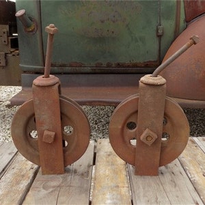 May include: Two rusty metal pulleys with a metal rod attached to each. The pulleys are mounted on a wooden surface.