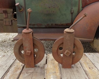 May include: Two rusty metal pulleys with a metal rod attached to each. The pulleys are mounted on a wooden surface.