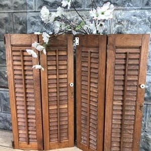 May include: Four wooden shutters with horizontal slats, arranged in a row. The shutters are a warm, medium brown color. White flowers are placed on top of the shutters. The shutters have small white knobs and hinges.