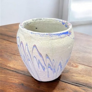 Könnte beinhalten: Eine cremefarbene Keramikvase mit strukturierter Oberfläche. Die Vase weist ein wellenförmiges Muster aus blauer, violetter und weißer Farbe auf. Der Rand der Vase ist ebenfalls mit blauen und violetten Akzenten bemalt. Die Vase steht auf einem Holztisch.