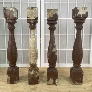 Könnte beinhalten: Vier antike Holzbaluster mit quadratischen Oberteilen und verzierten, gedrehten Körpern. Die Baluster sind in Braun- und Weißtönen lackiert, mit sichtbaren Abnutzungserscheinungen und Abplatzungen, die auf Alter und Gebrauch hindeuten. Sie stehen auf einer Holzoberfläche.