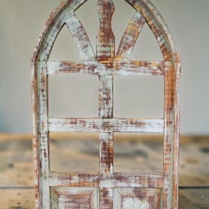 Puede incluir: Un marco de ventana decorativo de madera envejecida con una parte superior arqueada. El marco presenta un aspecto desgastado con tonos de marrón, blanco y azul pálido. El diseño incluye una viga vertical central y barras transversales horizontales.