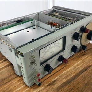 Puede incluir: Un analizador de distorsión Hewlett-Packard 334A vintage, un dispositivo electrónico gris con múltiples perillas, diales y un medidor. La parte superior abierta revela componentes internos. El dispositivo está sobre una superficie de madera.