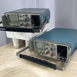 Puede incluir: Dos osciloscopios azules vintage con asas plateadas y paneles de control. Los osciloscopios tienen pantallas, perillas y botones. Uno está sobre un soporte blanco. Las asas son negras. Sobre una superficie de madera.