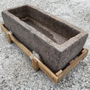 May include: A large, rectangular stone trough with a rough, textured surface. The trough is a muted gray color and sits on a simple wooden pallet. The trough's interior is hollow, suggesting it could be used for planting or as a decorative element.