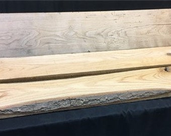May include: Two pieces of wood, one with a live edge and the other with a straight edge. Both pieces are light brown with a natural wood grain.