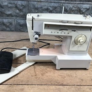 May include: A beige Singer sewing machine with a black foot pedal controller. The machine features a central dial for stitch selection, a needle assembly, and a flatbed base. The brand name Singer is printed in black letters on the front panel.