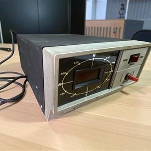 Puede incluir: Un dispositivo electrónico rectangular negro y plateado con una pantalla y un dial. El dispositivo tiene un interruptor rojo y un botón rojo. Un cable negro está conectado al dispositivo.