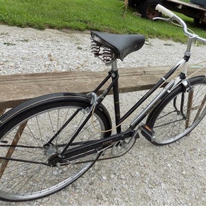 Op de afbeelding: Een vintage zwarte fiets met witte accenten, een zwart zadel en een verchroomd stuur. De fiets heeft een klassiek ontwerp met spatborden en een bagagerek. De banden zijn zwart met zilveren velgen. De fiets staat op een houten bank.