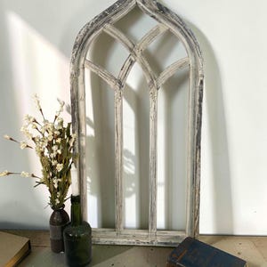 May include: A distressed, arched window frame with a weathered, off-white finish. The frame features a quatrefoil design at the top and vertical bars. A green bottle, a brown bottle, a candle, and a book are in front of the frame.