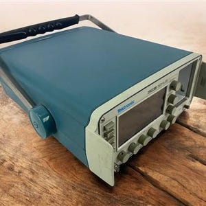 May include: A teal Tektronix 1503B signal generator with a black handle and silver frame. The front panel has a screen and several knobs. The device is on a wooden surface.