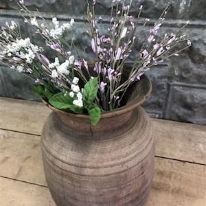 以下が含まれることがあります： 白と紫の花の茎と緑の葉で満たされた、素朴な茶色の木製花瓶。 花瓶は幅広で丸みを帯びたボディと、小さくフレアした上部を持っています。 背景には、質感のある灰色の石壁と木製の表面があります。
