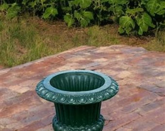 Puede incluir: Un macetero decorativo de hierro fundido de color verde oscuro con un diseño acanalado. El macetero está sentado en un patio de ladrillo frente a un campo de girasoles.