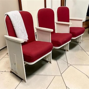 May include: A row of three theater seats featuring deep red fabric upholstery on the backrests and cushions, supported by white metal frames and armrests. A white patterned fabric cloth is draped over the backrest of the first seat.