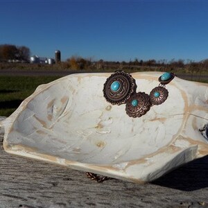 Puede incluir: Un plato de cerámica blanca con un acabado rústico y desgastado, que presenta un collar turquesa y cobre con un gran colgante circular.