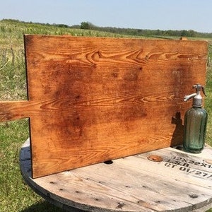 Puede incluir: Una tabla de cortar de madera grande, rectangular, con un asa en el lado izquierdo. La tabla está hecha de madera marrón claro y tiene un acabado natural y rústico. La tabla está sobre una superficie de madera.