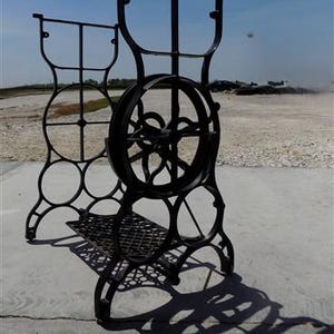 May include: Black antique sewing machine base with a large flywheel and decorative metalwork. The base features a grid-like platform and circular accents, casting a strong shadow on the concrete surface.