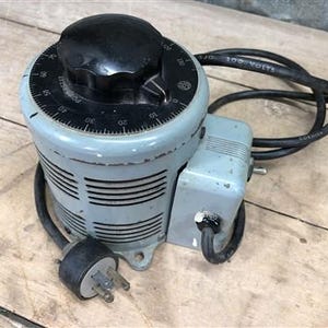 Könnte beinhalten: Ein grauer Variac-Autotransformator im Vintage-Stil mit einem schwarzen Knopf und Zifferblatt. Das Zifferblatt ist mit Zahlen und dem Wort "POWERSTAT" beschriftet. Er hat ein schwarzes Netzkabel und einen Stecker. Das Gerät steht auf einer Holzoberfläche.