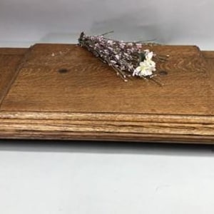 May include: A rectangular wooden board with a medium brown finish and visible wood grain, featuring a raised central panel with beveled edges. A small, decorative bundle of dried twigs with tiny white and pink flowers rests on the surface.