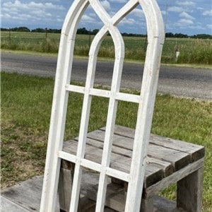 May include: A white, distressed-look decorative window frame with a gothic arch design. The frame has a grid of rectangular panes and is leaning against a rustic wooden bench outdoors. The sky is blue with scattered clouds.