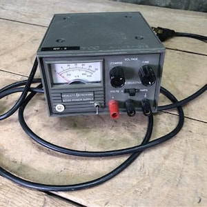 May include: A gray Hewlett Packard DC power supply with a black power cord. The front panel features a meter, knobs for voltage and current adjustment, and connection terminals. The meter displays readings in microamps. The power supply is on a wooden surface.