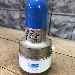 Puede incluir: Componente eléctrico cilíndrico con una parte superior azul, una sección media plateada y una base blanca. La base tiene una etiqueta con texto. El componente tiene una varilla metálica que se extiende desde la parte superior. Texto manuscrito visible.