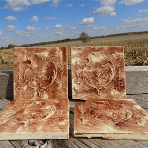 Può includere: Quattro piastrelle di ceramica antiche con un motivo floreale marrone e bianco. Le piastrelle sono quadrate e hanno un design in rilievo.