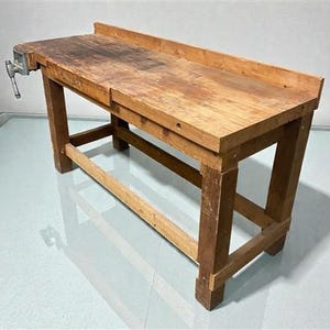 May include: A weathered wooden workbench with a metal vise attached to the left side. The workbench is constructed from light brown wood and features a raised back edge. The legs are sturdy and the overall design suggests a workshop setting.