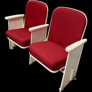 May include: Two theater seats featuring vibrant red fabric upholstery on the backrests and cushions. The chairs are supported by a sturdy white metal frame with matching white armrests on each side, showcasing a classic design for public seating.