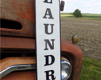 May include: White metal sign with black lettering that reads "LAUNDRY" in a vintage style font. The sign is attached to a rusty truck.