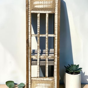 Puede incluir: Una pieza de decoración de pared rectangular y alta de madera con un acabado desgastado en marrón y blanco. El marco tiene una estructura de rejilla con listones verticales y horizontales, similar a una ventana, con paneles sólidos.