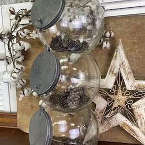 May include: Three glass jars with metal lids stacked on top of each other. The jars are filled with pine cones and have a rustic farmhouse style.