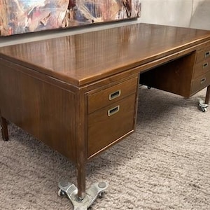 May include: A large, vintage wooden desk with a glossy finish. The desk has multiple drawers with brass-colored handles and sits on caster wheels. The desk is approximately 60 inches wide and 30 inches deep.