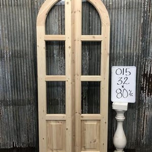 May include: A pair of unfinished wooden arched doors with glass panes. The doors have a light wood grain and are displayed against a corrugated metal background. A sign next to the doors reads "015 x 32 x 80 1/2".