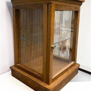 May include: A wooden display cabinet with a glass door and shelves. The cabinet is made of a medium-toned wood and has a rectangular shape. The door has a small brass knob. The cabinet is designed to showcase collectibles.