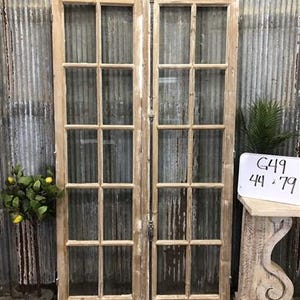 May include: A pair of weathered, light-colored wooden doors with multiple glass panes. The doors are propped against a corrugated metal wall. A small sign with the numbers "44 x 79" is visible. A decorative plant is to the left.