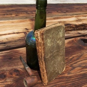 Puede incluir: Una botella de vidrio verde con un libro encuadernado en cuero marrón y una pipa de madera sobre una superficie de madera.