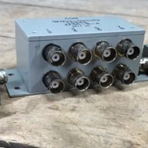 Può includere: Tre componenti elettronici grigi con più connettori BNC. Il componente più grande ha otto connettori, mentre gli altri due ne hanno uno ciascuno. Ogni componente ha del testo stampato.