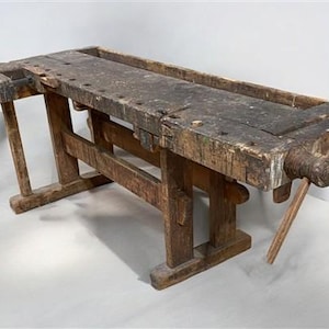 May include: A weathered, antique wooden workbench with a dark, aged surface. The workbench features a long, rectangular top with a built-in trough and a vise on one end. The legs and frame are also made of wood, showcasing a rustic, vintage aesthetic.