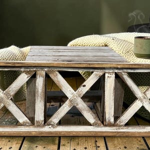 May include: A rustic wooden coffee table with a distressed white and brown finish. The table features an X-shaped design on the sides and a slatted top. A green mug and cream-colored blanket rest on top, creating a cozy, farmhouse aesthetic.