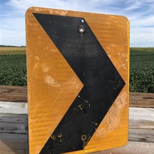 May include: A yellow and black road sign with a black arrow pointing to the left. The sign is made of metal and has a reflective surface.