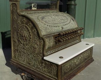 Könnte beinhalten: Antike Messing-Kasse mit aufwendigen floralen Details. Sie verfügt über eine weiße Marmorablage und eine Glasanzeige mit der Zahl "40". Die Kasse hat mehrere Tasten und ein dekoratives Design.
