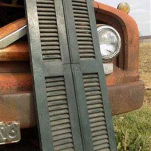 May include: A pair of antique wooden shutters painted in a dark green color. The shutters are closed and have a weathered look. They are approximately 75 inches tall and 20 inches wide.