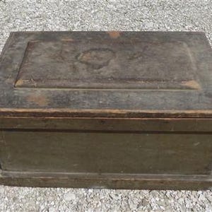May include: A rectangular, antique wooden box with a hinged lid. The box is painted in shades of green and brown, with visible wear and tear, suggesting age. The lid features a recessed panel and a decorative element. The box sits on a gravel surface.